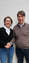 To personer smiler og håndhilser foran en lys vegg.