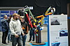 Flere besøkende står foran en fargerik rørinstallasjon og informasjonsplakater på en messe.