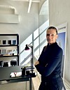 Person ved skrivebord i lyst kontor med hyller, lampe og sollys som faller inn gjennom vinduet.